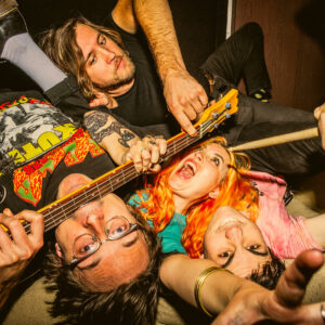 The four band members in something of a pile, in a photo shot from above them, looking up with a bass guitar and a drumstick in hands, and smiles on faces.