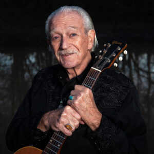 Charlie Musselwhite holding an acoustic guitar, wearing a black Western shirt.
