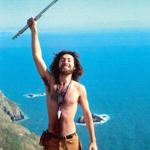 Iasos holding a flute triumphantly on top of a mountain with the ocean in the background.