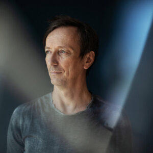 Profile picture of Volker Bertelmann, aka Hauschka, with a black and blue background.