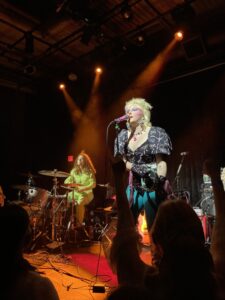 Caroline Kingsbury singing, head tilted up, into a microphone on stage at Rickshaw Stop with their drummer in the background