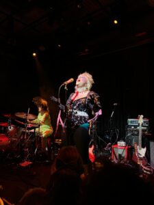 Caroline Kingsbury singing, head tilted up, into a microphone on stage at Rickshaw Stop with their drummer in the background