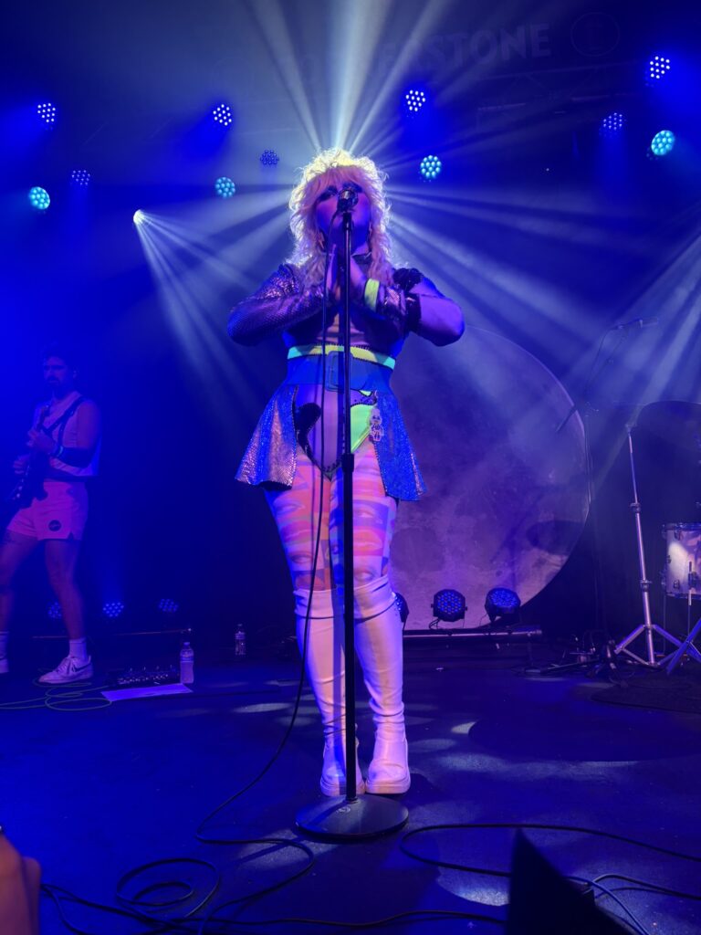 Caroline Kingsbury standing up straight at the mic stand arms folded with a beam of stage lighting coming from behind