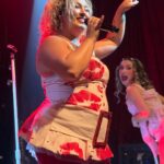 Smiling Molly Grace, hands up and holding a mic with one backup singer turned away from the audience mouth open